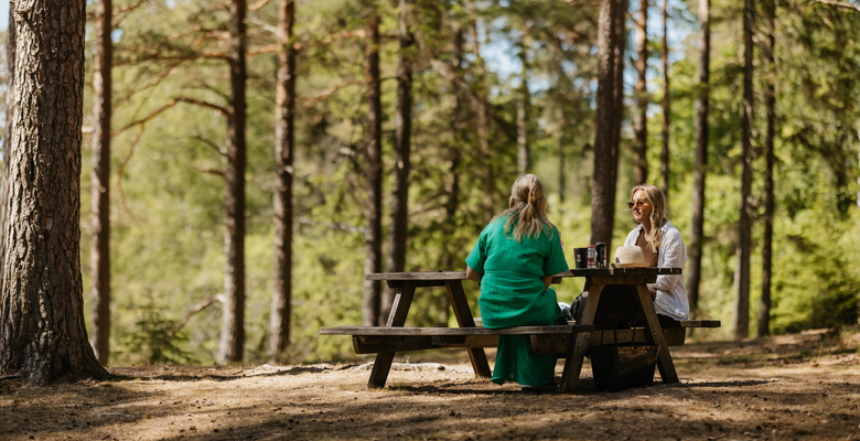 Två personer sitter på en bänk i skogen och kollar på varandra. På bordet står vattenflaskor
