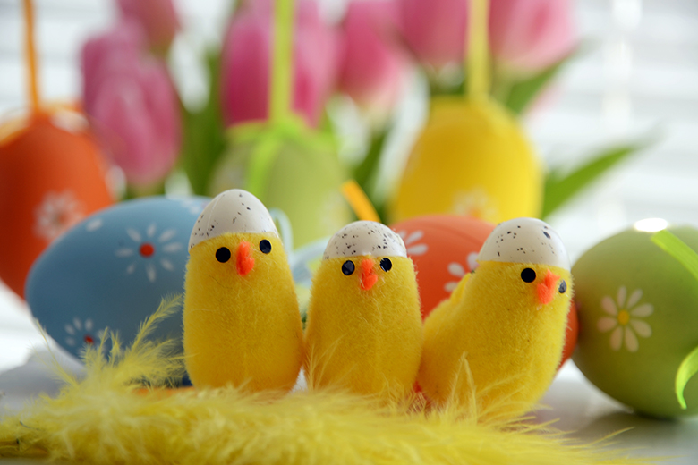Tre gula påskkycklingar i tyg som sitter framför färgglada ägg
