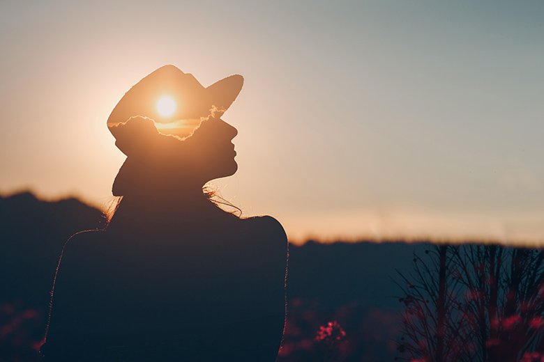 Kvinna som står vid en soluppgång. Hon bär en hatt där solen lyser igenom. 